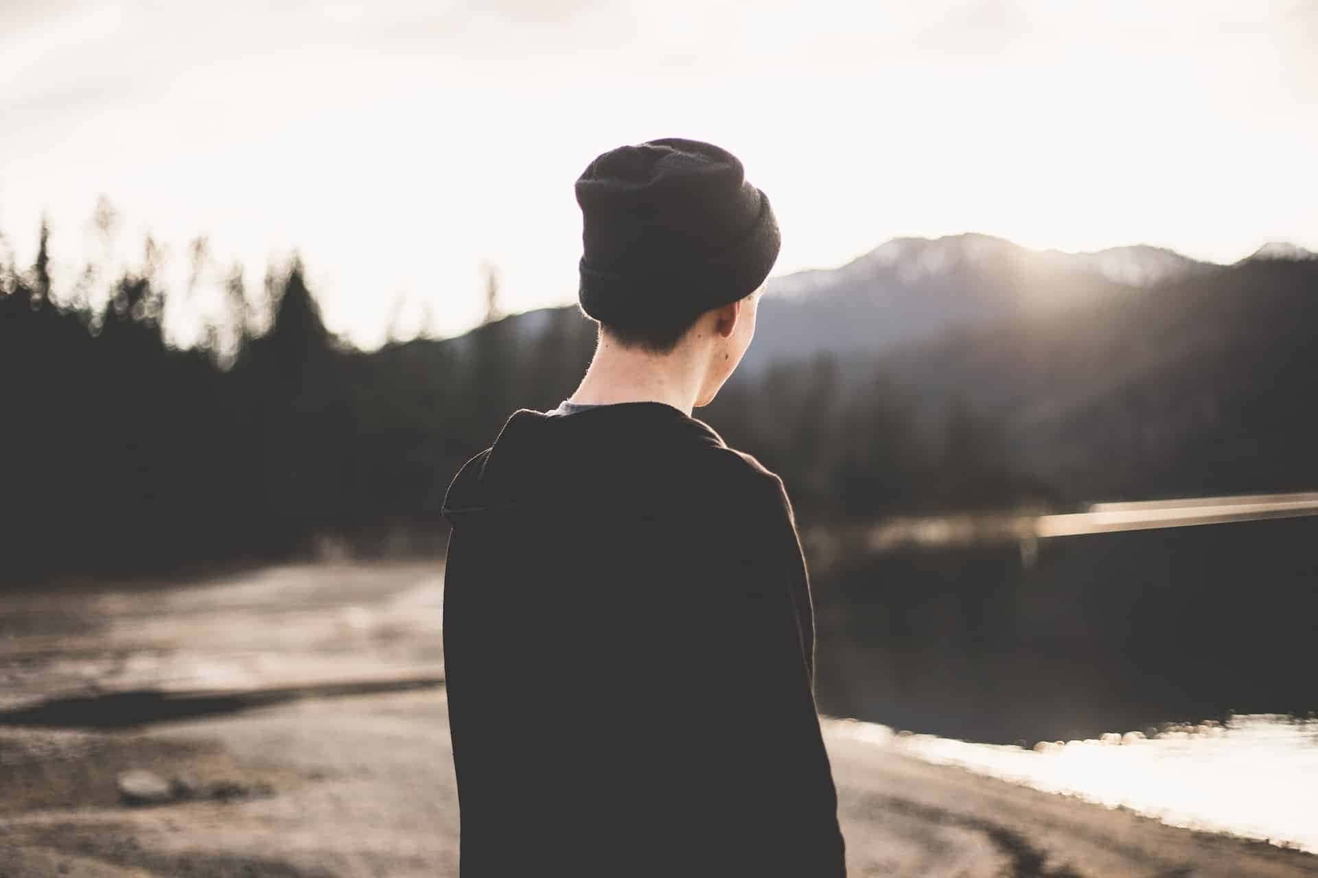 a man wearing a black beanie is standing by a lake.