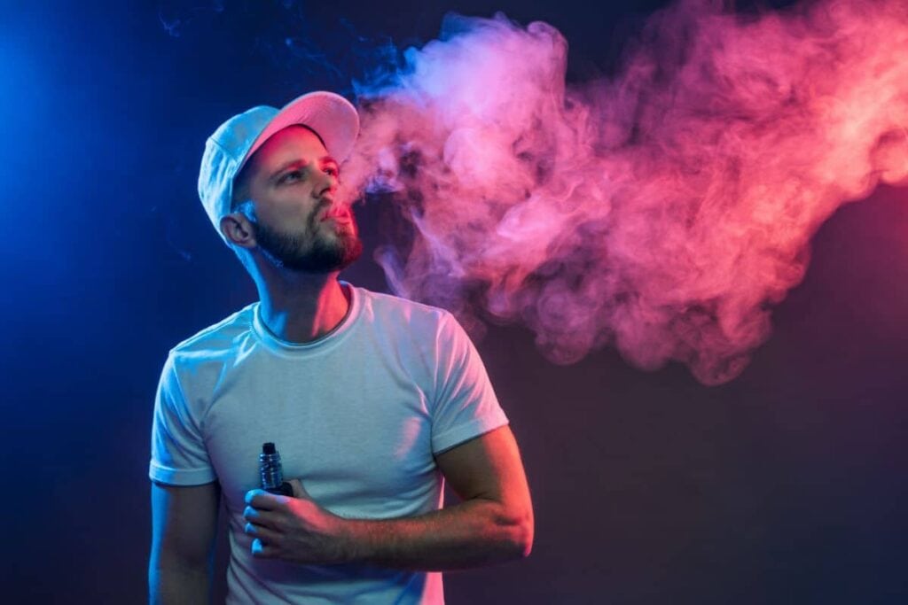 Man exhaling vapor from a do thca electronic cigarette against a colorful background.