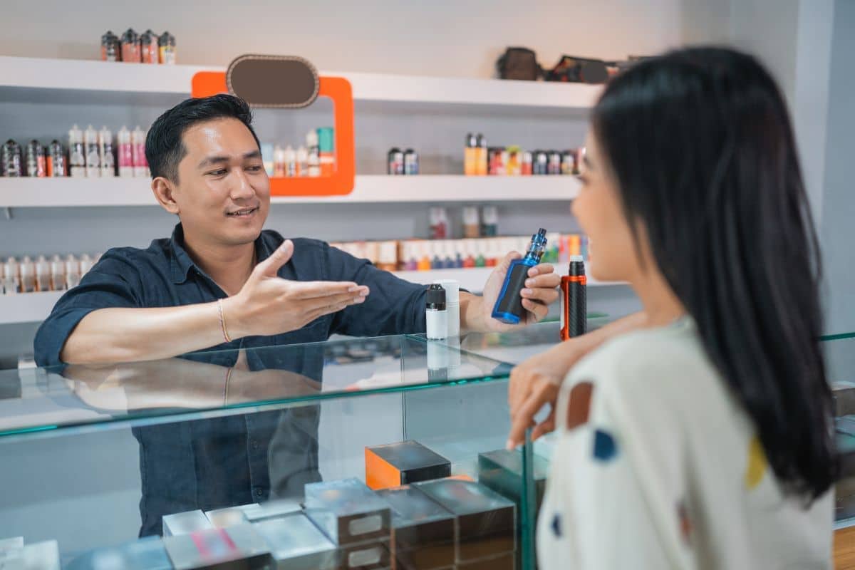 Salesperson explaining mailing regulations for e-cigarettes to a female customer at a vape shop.